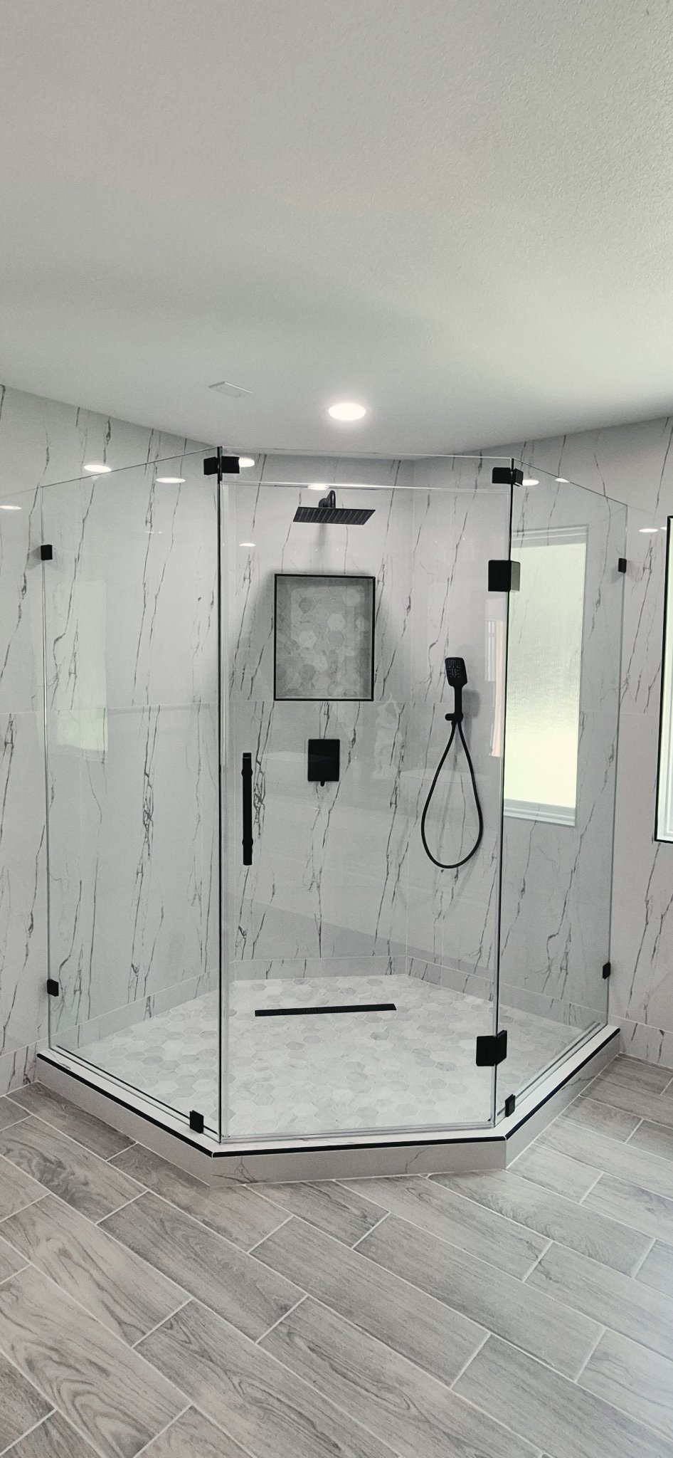 Foto de referencia del baño en ángulo 135° (neo-angle): shower de vidrio en esquina con tres lados, puerta central y herraje negro, en un baño con paredes de mármol
