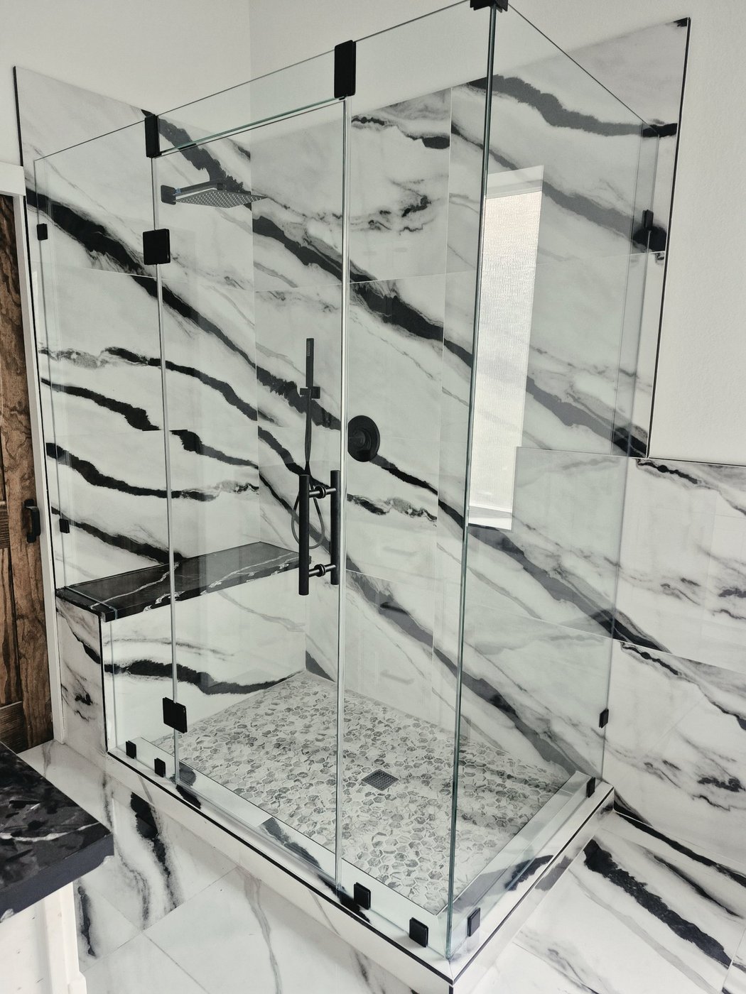 Trabajo de Glas Plus: ángulo 90° con herraje negro en un baño de mármol con vetas negras y banca interior