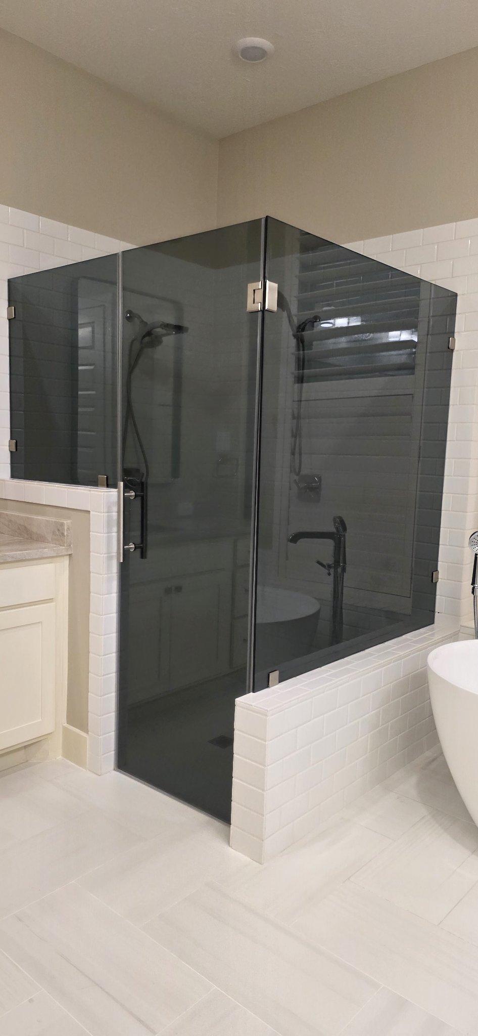 Trabajo de Glas Plus: shower en ángulo 90° con vidrio Grey (gris ahumado) montado sobre muros de azulejo blanco, junto a una tina