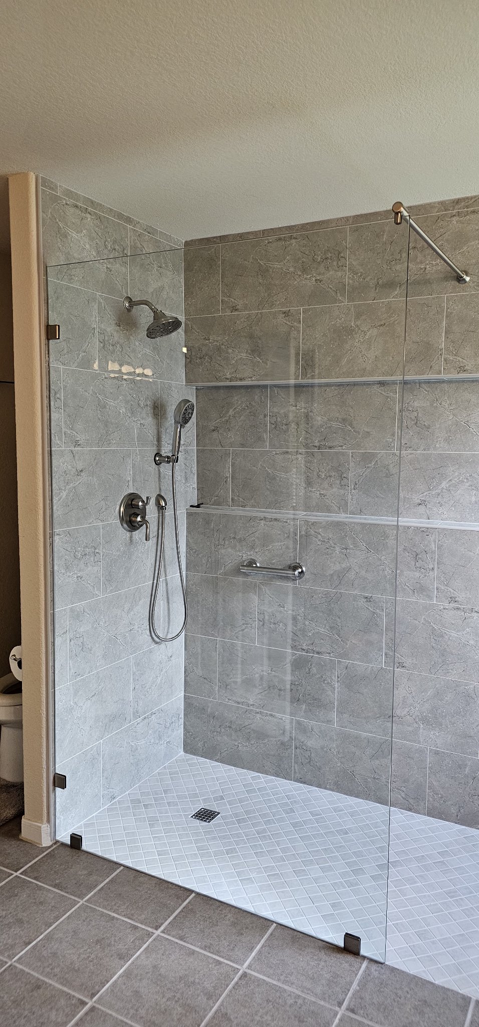 Foto de referencia del panel fijo ancho de Glas Plus: un panel fijo de vidrio amplio, sin puerta, en un shower abierto de azulejo gris, con una barra estabilizadora diagonal en la parte superior