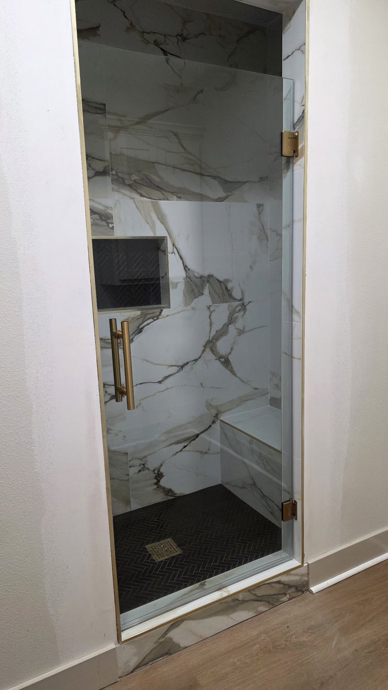 Foto de referencia de la single door de Glas Plus: una sola puerta de vidrio de bisagras, sin panel, con herraje brush gold en un shower de mármol