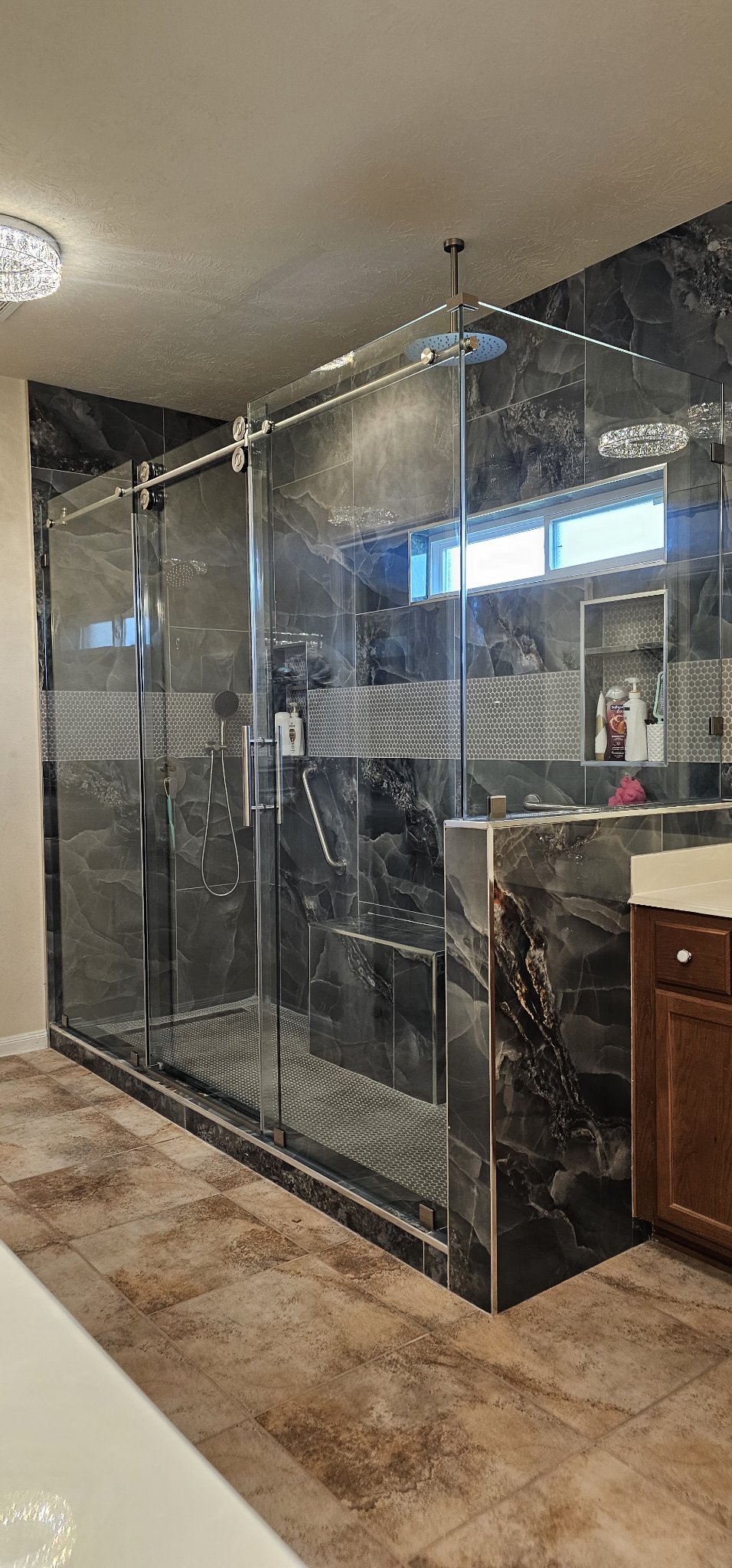 Ejemplo de sliding de una puerta con panel fijo de Glas Plus: puerta corrediza de vidrio sobre riel cromado junto a un panel fijo, en un baño amplio de mármol oscuro